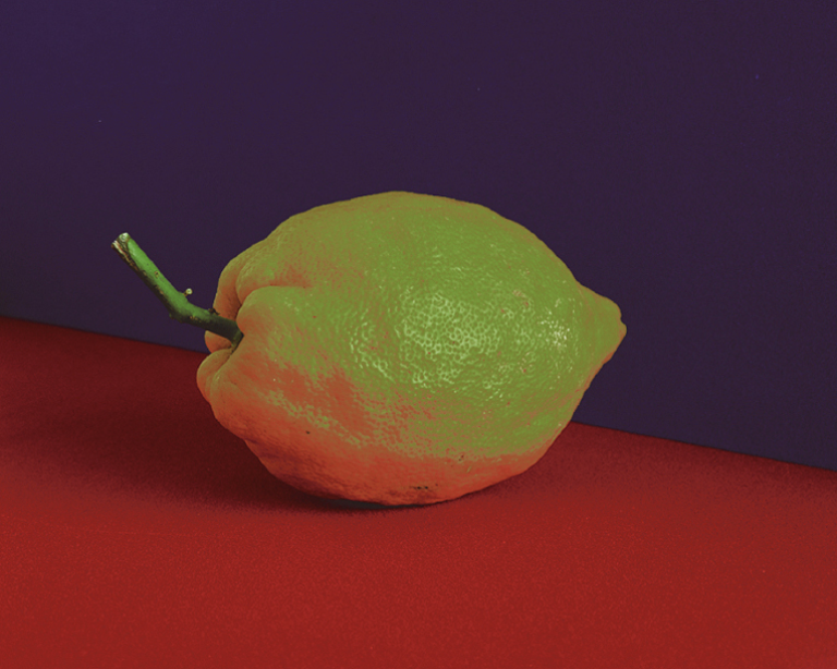 A lemon with its stem attached sitting on a flat surface. The image is recolored such that the lemon appears more green than yellow, and the lower part that would typically be gray with shadow is an orangey-red. The surface below it is red, and the background color is a deep purple. Carina Lewandowski