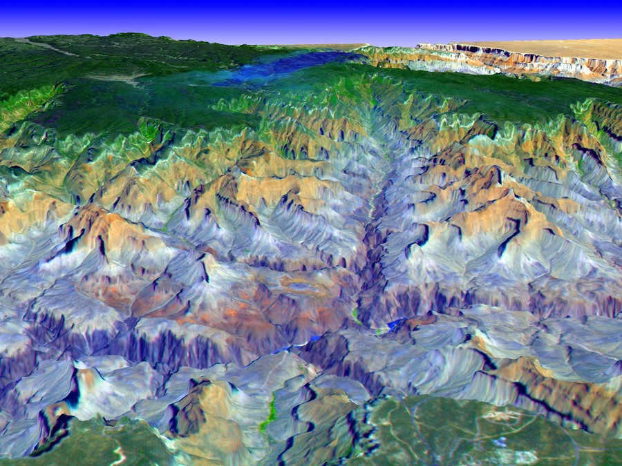 The image is an aerial photograph of the Grand Canyon. The horizon line is near the top of the composition, above which a blue sky is seen. The canyon is below the horizon line. Deep crevices filled with water run across and through the canyon, which recedes straight toward the vanishing point at the top-center of the composition. The image is colored unnaturally with blue, green, and beige tones to help the viewer delineate the mountainous forms within the canyon; specifically, the shadows are often described by a saturated dark blue to draw out the various elevation changes, while the peaks of the inter-canyon ridges are light-beige and off-white. Drew Pugliese