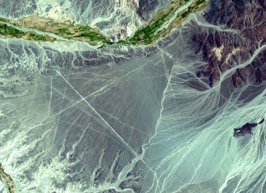 The image is an aerial photograph of the Nazca lines in Peru. The composition includes rivers, mountains, and forests, but is primarily filled with a large desert. Criss-crossing the desert are thin, straight white lines that exist in contrast to the other organic, non-linear geological forms. The image is colorized unnaturally using green, blue, purple, white, and black tones. Drew Pugliese
