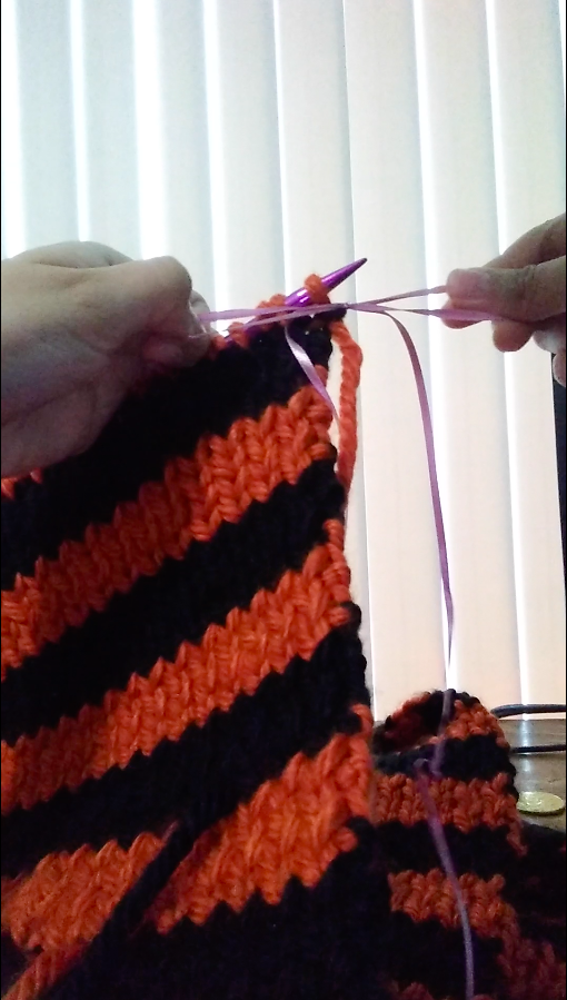 Close-up image of two hands tying a bow around a stitch in the top row of a work-in-progress knit scarf. The scarf has orange and black stripes. The bow is being created using a thin pink string. This image is a screenshot from a video of me knitting. Jess Ramirez