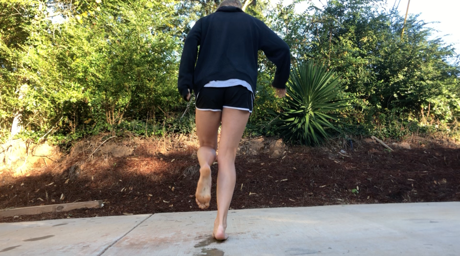A human is pictured hoping on her right foot on a concrete driveway from behind. The background is a progression from the gray driveway to the dirt underneath the lush green plants beneath the blue, bright sky. The hopper is upright, but the driveway slants down to the left. Footprints mark the previous path of the hopper. Kara Steele