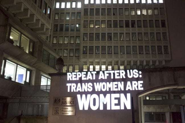 The text “Repeat after us: trans women are women” projected in white, capital block letters on the Ministry of Justice building in London to raise awareness about the injustices faced by the trans community. To the left of the text are two silver plates; one says “Ministry of Justice,” and the other says “Crown Prosecution Service.” There are other office buildings covered in windows to the left and behind the building where the text is projected. Most of these windows are dark with the exception of the row of windows at the very top of the image. Carina Lewandowski