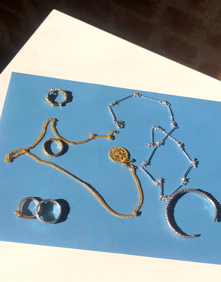 The image shows a picture of a blue piece of paper resting on the corner of a white table. On top of the paper are several pieces of gold and silver jewelry. In the bottom-left corner of the paper are two overlapping rings-one gold, and one silver. In the bottom-right corner is a silver cuff. In the top-left corner are two gold rings. Draped across the paper are one gold and one silver necklace. Drew Pugliese