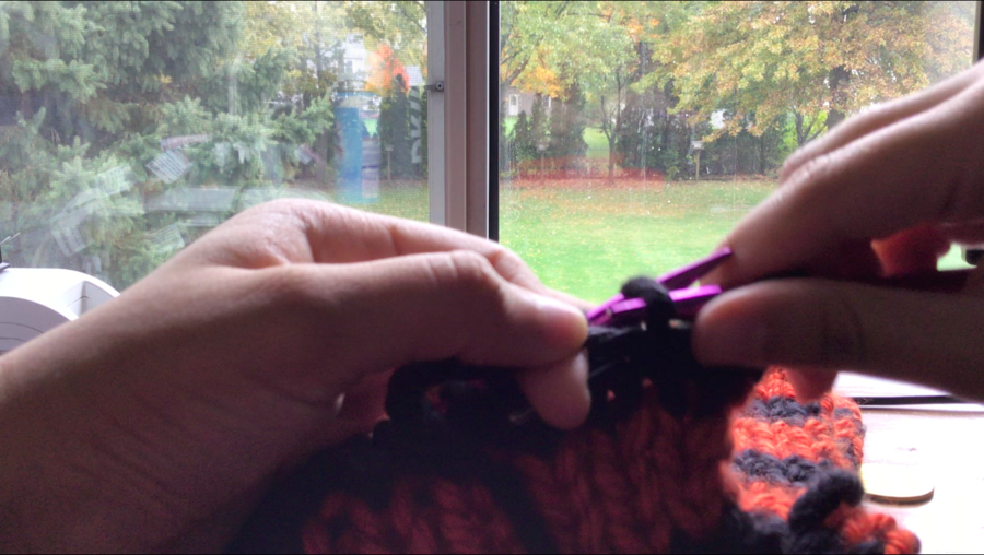 Close-up image of two hands knitting an orange and black striped scarf. Green trees and grass can be seen through a window in the background. This image is a screenshot from a video of me knitting. Jess Ramirez