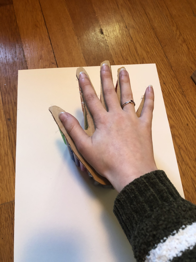 The image shows a hand with a ring on the ring finger resting on top of a piece of cardboard cut into the shape of a hand. Below the cardboard is a white piece of paper on a wooden floor. The arm of the hand extends off the bottom right corner of the photo and is wearing the sleeve of a green and white striped sweater. Katie Miller