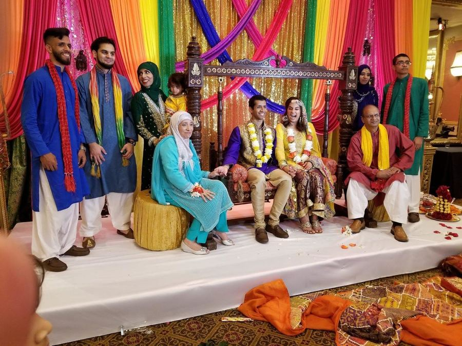 Picture of my family at my brother's mehndi. A pakistani family dressed in traditional pakistani shalwar is standing around a bride and groom. Everyone is wearing very colorful clothing that matches the background of pink, purple, green, blue, orange, and golden. The couple has flowers draped around their neck and the grooms parents are on either side of them. The grooms siblings are next to the parents. Sameer Riaz