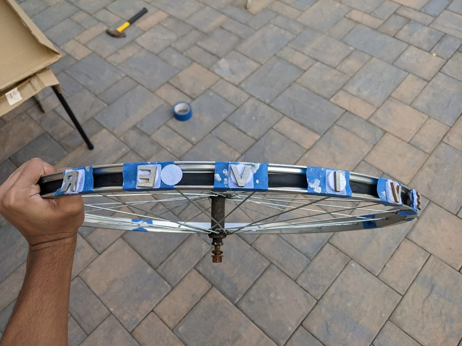 A lone bicycle rim with pieces of tape placed over the gapes and wooden letters attached to the tape with velcro. The letters are covered in silver paint. A brown hand can be seen holding the rim in the air over a brick floor, and a roll of blue electrical tape and a hammer can be seen on the floor. Sameer Riaz