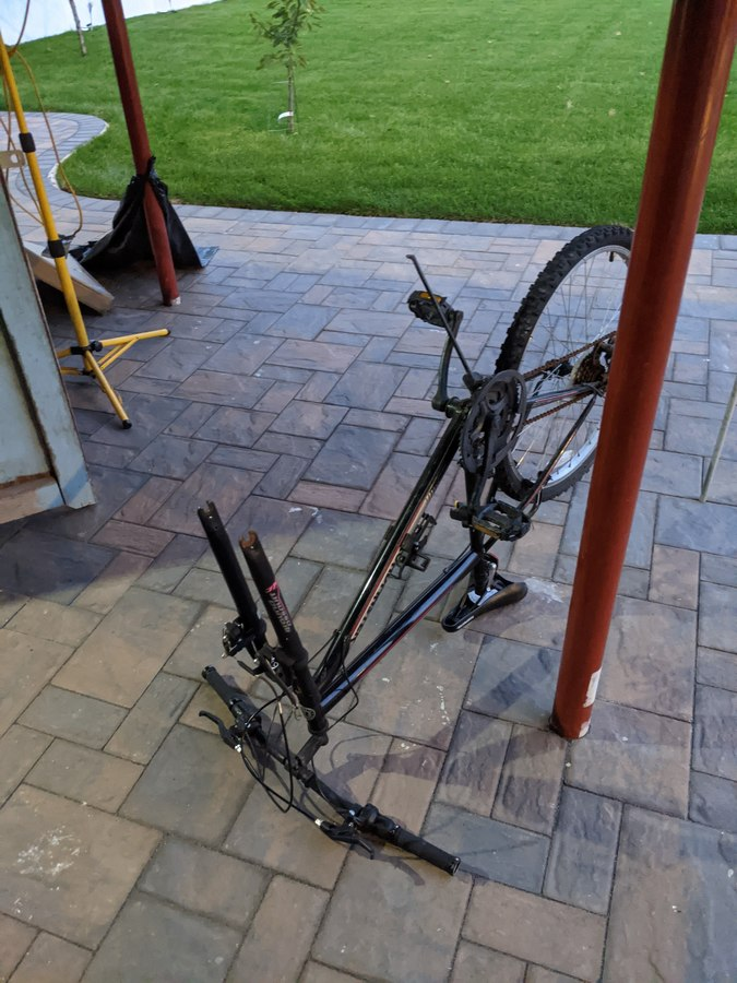 A black bicycle with the front wheel removed from it resting on a brick patio in a backyard. The grass is visible in the back of the photo, and there are red support poles in the picture. Sameer Riaz