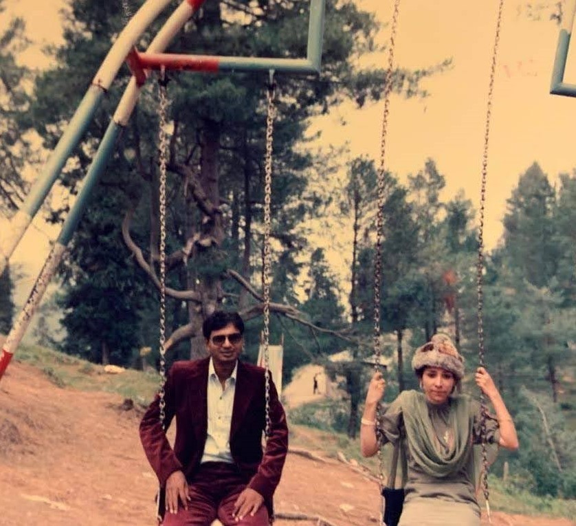 A young, newly married Pakistani couple on their honeymoon sitting on a swingset in a northern wooded region of Pakistan. The man is wearing sunglasses and a maroon felt suit and a white shirt, and the woman is wearing a mint shirt and dupatta with a gray fur hat. Sameer Riaz