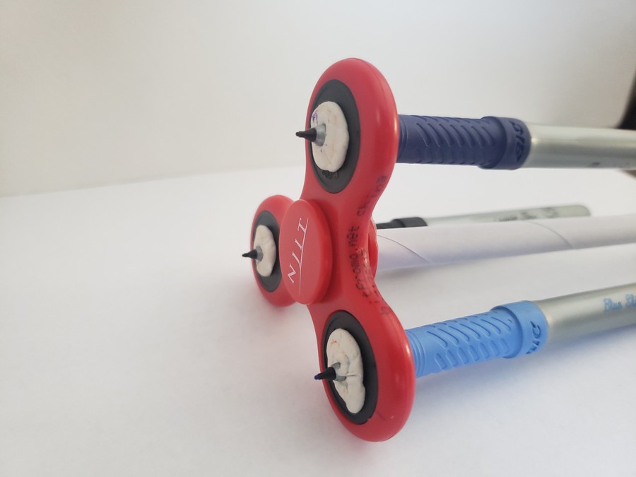 A fidget spinner printer composed of three colored markers attached to the holes of a red NJIT fidget spinner with white puddy. The markers are attached to the underside of the spinner such that their nibs face left and their tops are off to the right. In addition to the markers, a white paper rod extends from the center of the fidget spinner to the right. Thanya Begum